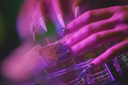 Close-up of hands with MiuNail Venom Twist black and white nail wraps holding a cocktail glass under moody purple and green lighting.