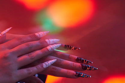 Hands wearing MiuNail Venom Twist black and white sculpted nails, under vivid multicolor lighting with red and orange glow.