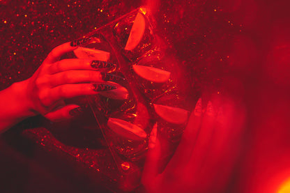 Hands with MiuNail Venom Twist black and white nails holding a cocktail tray under red lighting, surrounded by sliced citrus and shimmering glitter.