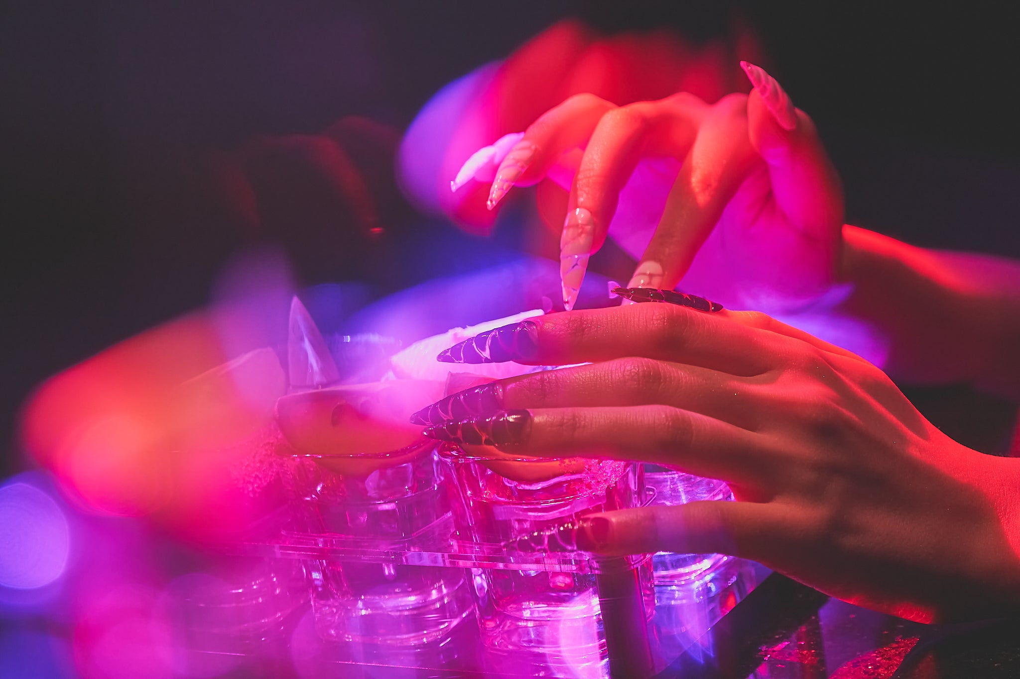 Close-up of hands with MiuNail Venom Twist nails reaching over cocktail glasses, illuminated by vivid pink and purple lighting.