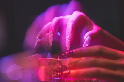Close-up of hands wearing MiuNail Venom Twist white sculpted nails, holding a cocktail under pink and purple neon lighting.