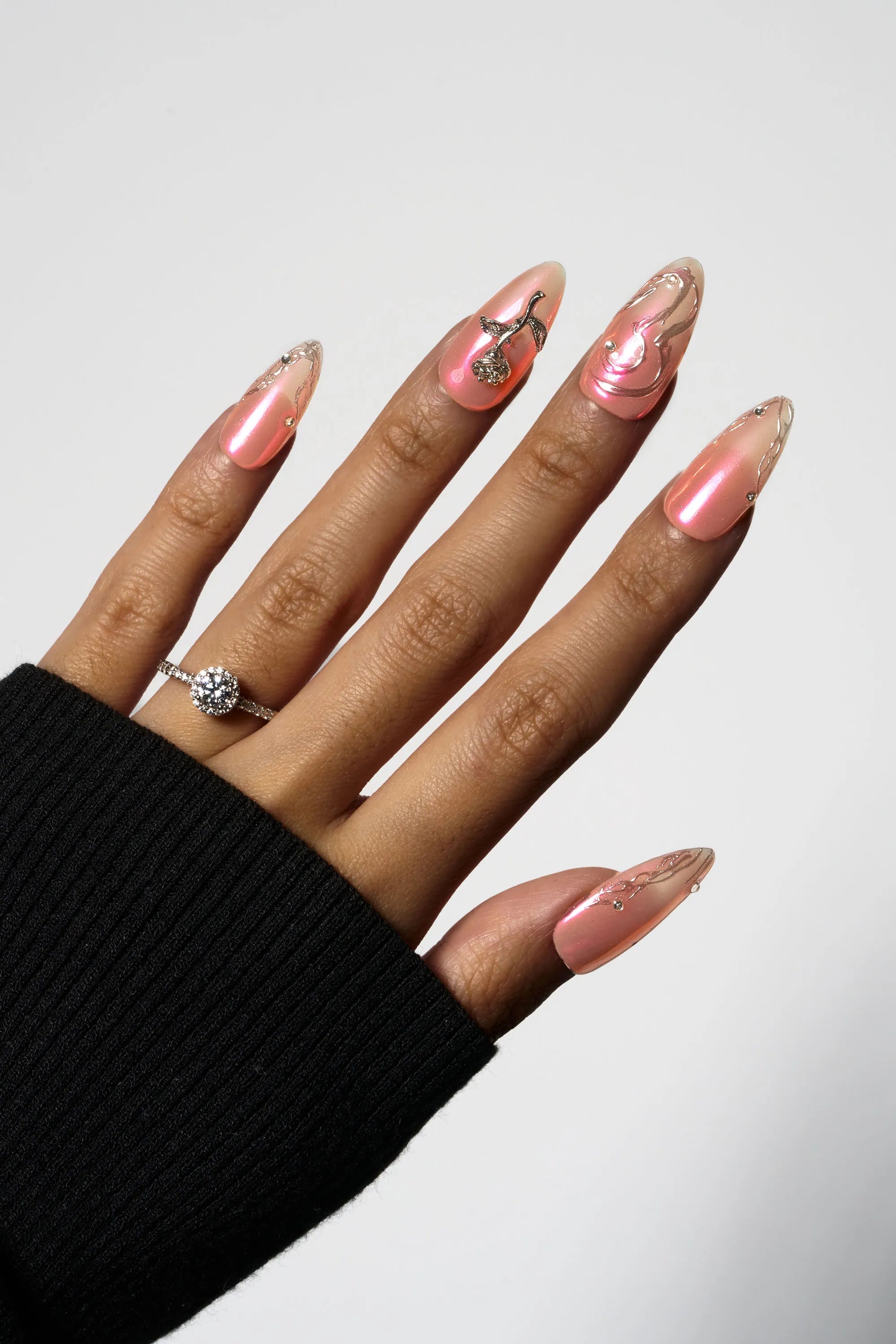 Molten Coral almond-shaped press-on nails with a pearlescent shimmering pink finish, featuring a prominent 3D metallic gold rose charm and liquid gold swirl accents.