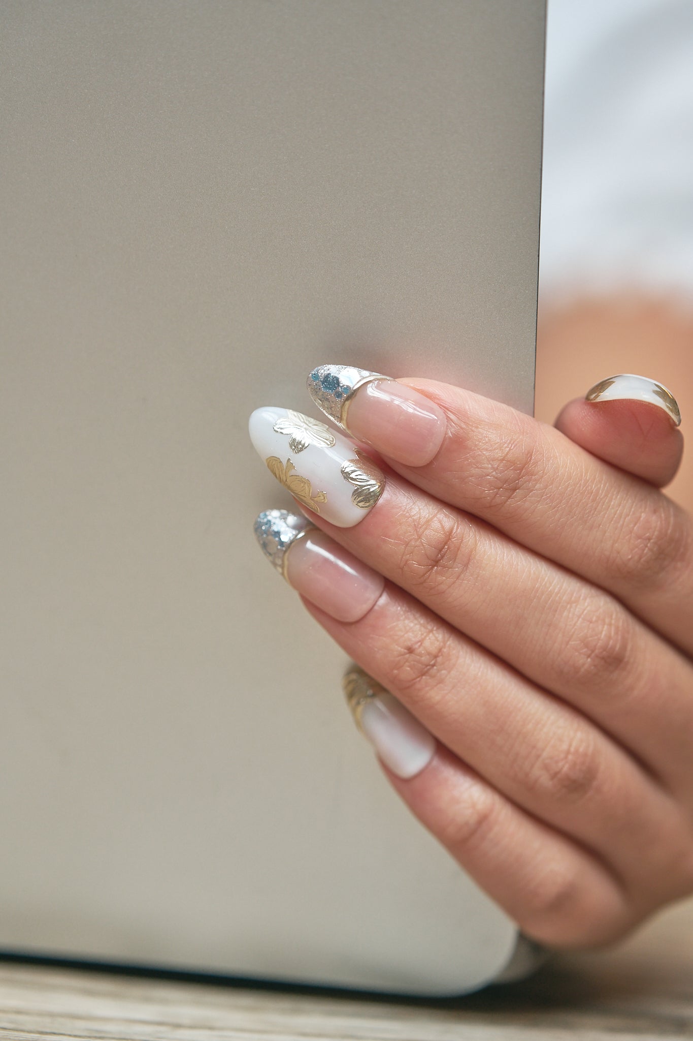 Close-up of a hand adorned with Petal Bloom nails holding a laptop against a soft neutral background, highlighting the intricate gold and silver nail art.