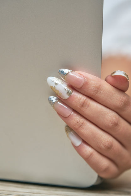 Close-up of a hand adorned with Petal Bloom nails holding a laptop against a soft neutral background, highlighting the intricate gold and silver nail art.