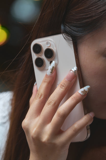 Close-up of a hand with Petal Bloom nails holding a smartphone to the ear, showcasing intricate gold and silver nail designs with a blurred background.