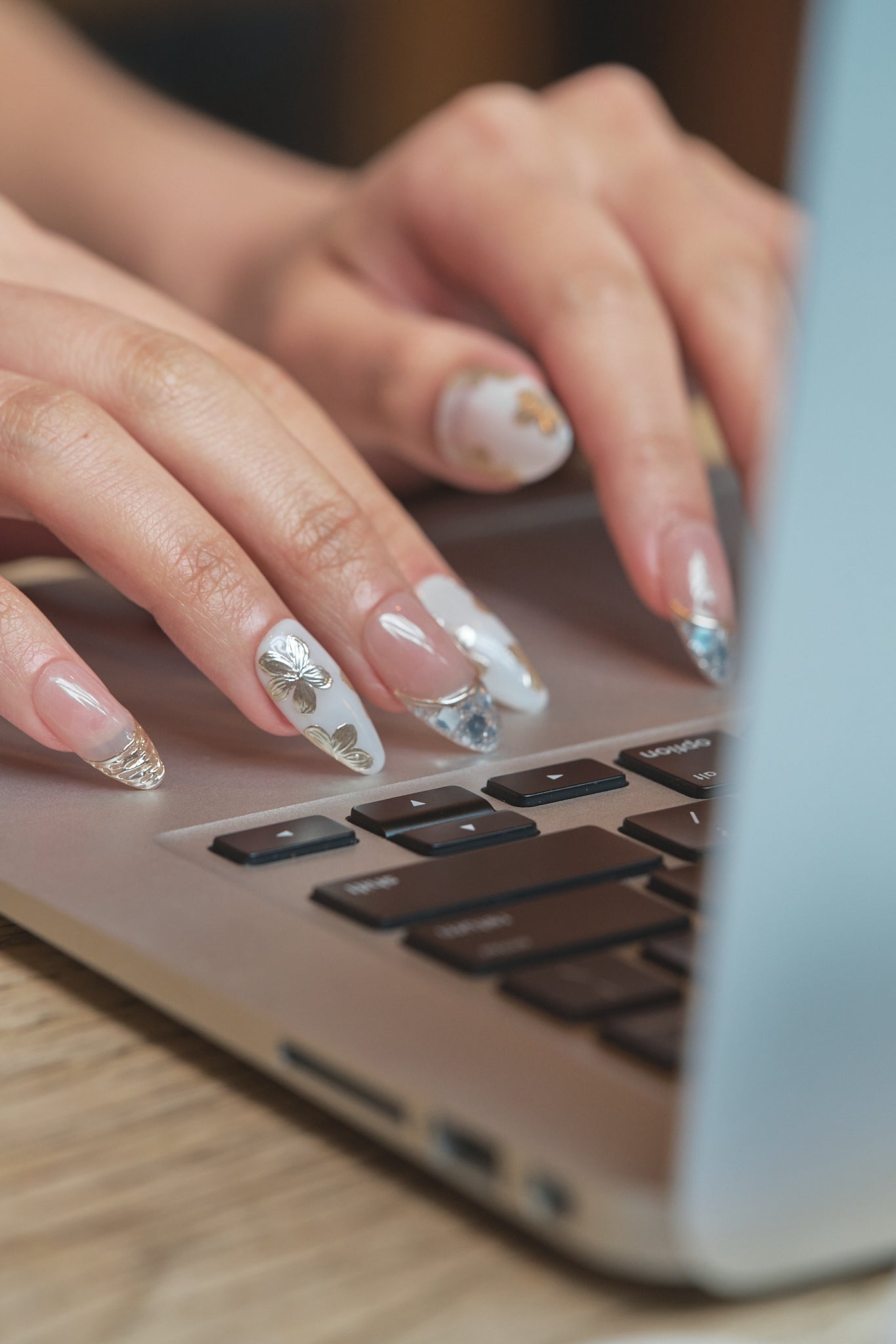 Close-up of hands with Petal Bloom decorative nails typing on a laptop keyboard, highlighting delicate gold and silver nail art.