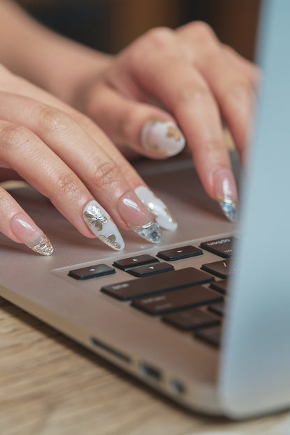 Close-up of hands with Petal Bloom decorative nails typing on a laptop keyboard, highlighting delicate gold and silver nail art.