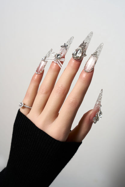 Rosé Glacé reusable extra-long stiletto press-on nails showcasing a sheer pink cat-eye shimmer, decorated with delicate white ribbon bows, 3D silver heart charms, and dangling crystal teardrop accents.