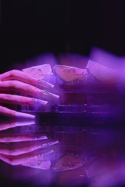 close-up of a hand with long, rhinestone-decorated nails resting on a reflective surface with lemon slices and purple neon lighting